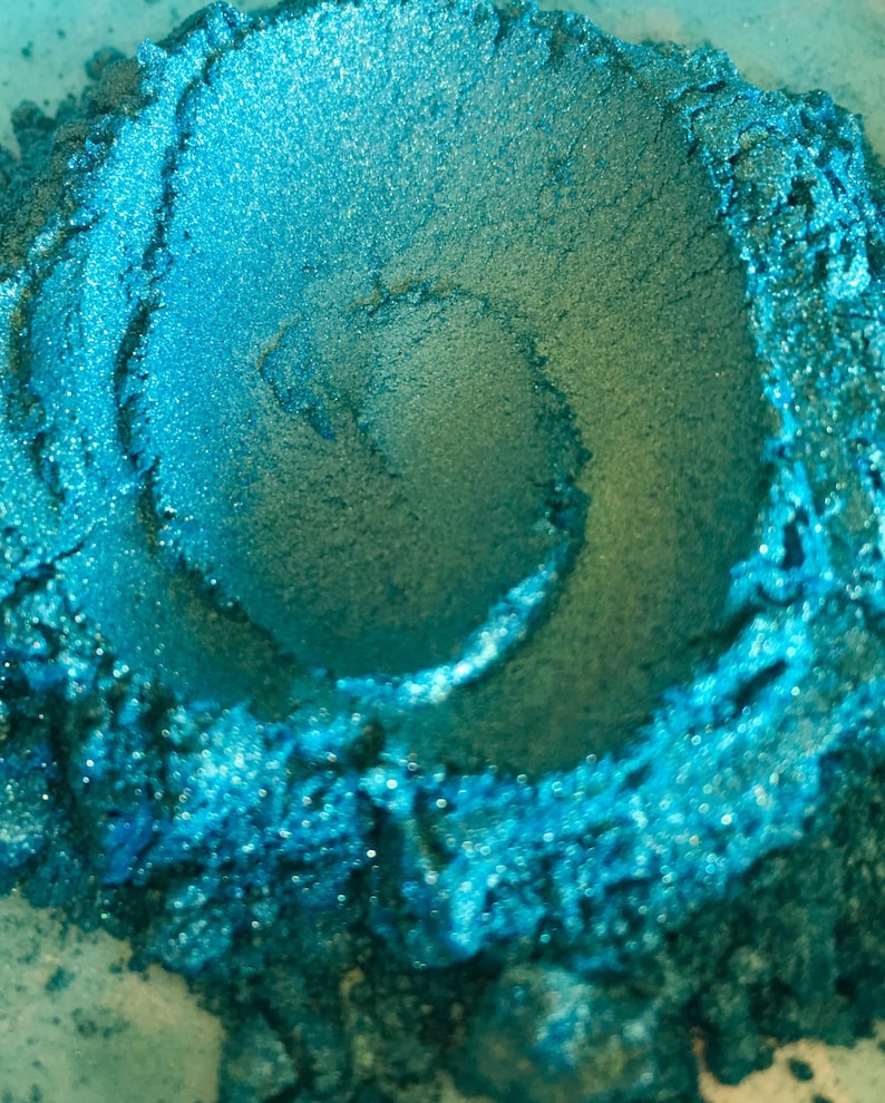 May include: Close-up of a pile of teal-colored pigment powder. The powder has a shimmering, metallic finish and a swirling pattern in the center. The texture appears fine and granular, with variations in color and depth.