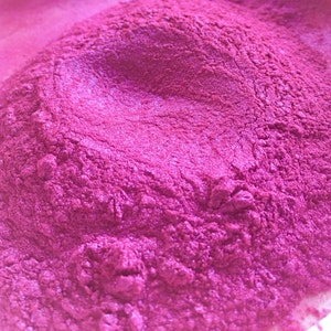 May include: Close-up of a pile of vibrant, magenta-colored powder with a shimmering, metallic finish. The powder has a fine, textured appearance, with subtle variations in tone and highlights. The image suggests a cosmetic or craft supply.