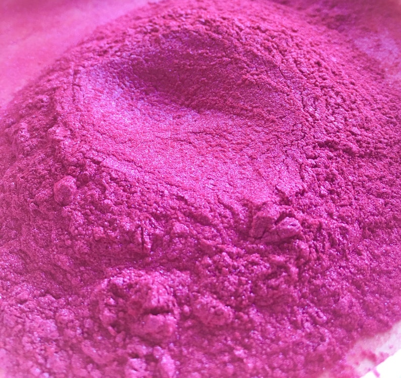 May include: Close-up of a pile of vibrant, magenta-colored powder with a shimmering, metallic finish. The powder has a fine, textured appearance, with subtle variations in tone and highlights. The image suggests a cosmetic or craft supply.