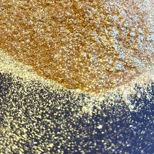 Op de afbeelding: Close-up van een oppervlak bedekt met gouden glitter. De glitter is dicht opeengepakt, waardoor een textuur ontstaat. Het bovenste gedeelte is scherp gefocust, terwijl de onderste helft een wazig effect heeft, met een donkerblauwe achtergrond.