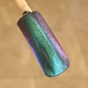 May include: A nail swatch displays a shimmering, iridescent polish. The color shifts from blue-green to purple, with a glittery finish. The sample is on a white stick, showcasing the color's vibrancy.