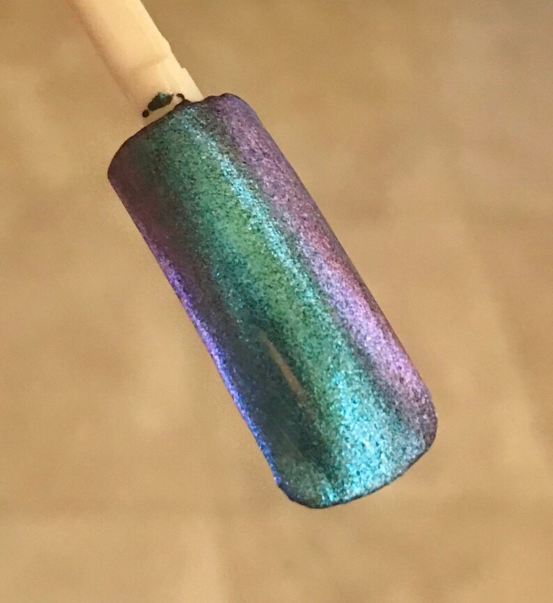 May include: A nail swatch displays a shimmering, iridescent polish. The color shifts from blue-green to purple, with a glittery finish. The sample is on a white stick, showcasing the color's vibrancy.
