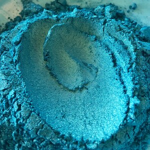 May include: Close-up of a pile of teal-colored powder with a metallic sheen. The powder has a textured, granular appearance, with areas of light and shadow. The overall effect is a shimmering, iridescent teal. This could be a pigment or cosmetic product.
