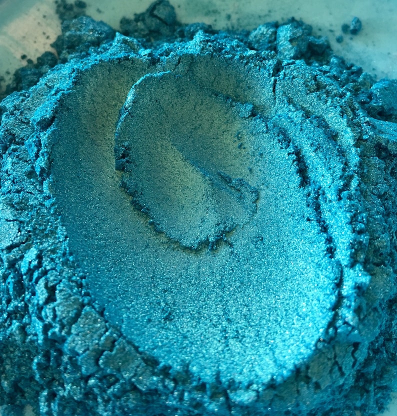 May include: Close-up of a pile of teal-colored powder with a metallic sheen. The powder has a textured, granular appearance, with areas of light and shadow. The overall effect is a shimmering, iridescent teal. This could be a pigment or cosmetic product.