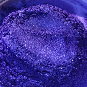 May include: Close-up of a pile of vibrant, shimmering purple pigment powder. The powder has a textured appearance with visible glitter particles, creating a sparkling effect. The color transitions from deep violet to lighter shades.