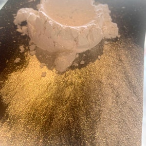 May include: A pile of white, shimmering powder sits on a dark, reflective surface. The powder has a textured, granular appearance, with visible sparkles. The surface below has a golden, glittery sheen, creating a contrast.