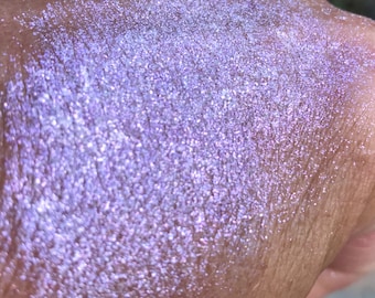 May include: Close-up of a hand dusted with iridescent, shimmering powder. The powder has a lavender and blue color with a sparkling, glittery effect. The texture appears smooth and finely milled, reflecting light.