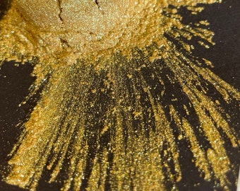 May include: Close-up of a golden powder with a shimmering effect, spread across a black surface. The powder is concentrated in a circular shape, with radiating lines of glitter extending outwards, creating a burst-like pattern.
