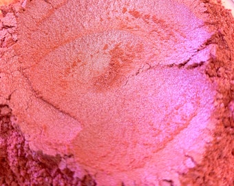 May include: Close-up of a container filled with a shimmering, reddish-pink powder. The powder has a metallic sheen and appears finely milled, with subtle variations in color and texture. The overall effect is a vibrant, iridescent pigment.