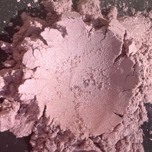 Pode incluir: Close-up de um monte de pó brilhante, rosa malva claro. O pó tem uma superfície texturizada com rachaduras visíveis e um brilho sutil. A cor é um rosa suave e suave com um brilho perolado, sugerindo um produto cosmético.