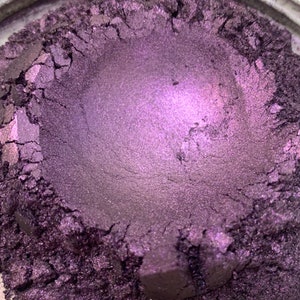 May include: Close-up of a container filled with a shimmering, deep purple powder. The powder has a metallic sheen and a textured surface, with visible cracks and crevices. The color transitions from a darker shade to a lighter, almost lavender hue in the center.