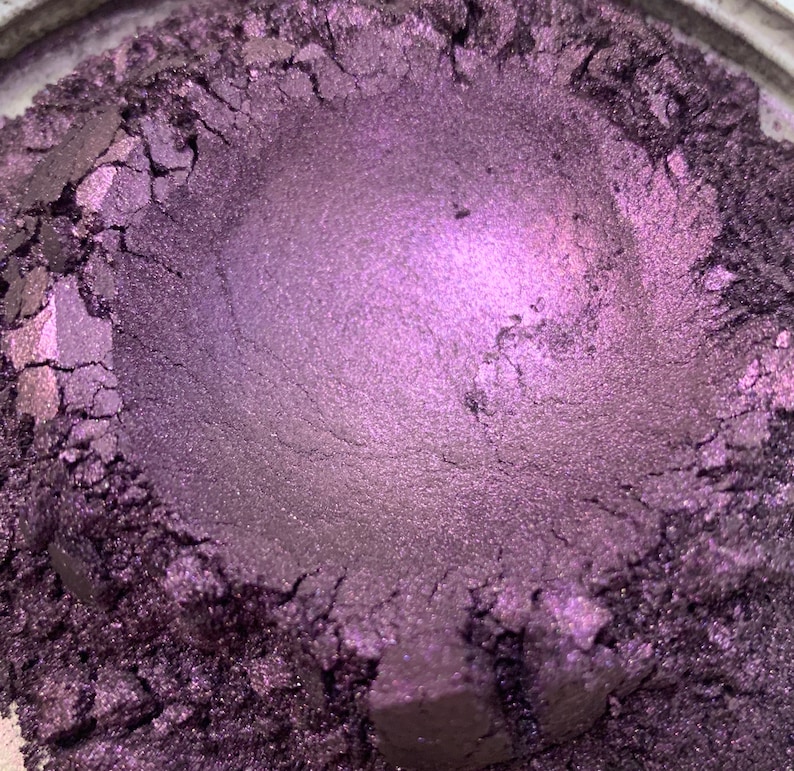 May include: Close-up of a container filled with a shimmering, deep purple powder. The powder has a metallic sheen and a textured surface, with visible cracks and crevices. The color transitions from a darker shade to a lighter, almost lavender hue in the center.