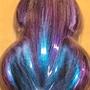 May include: A teardrop-shaped object with a shimmering, iridescent finish. The colours shift from deep purple to vibrant blue and teal, creating a glossy effect. The surface reflects light, highlighting the texture. The object is approximately 10 cm in length.