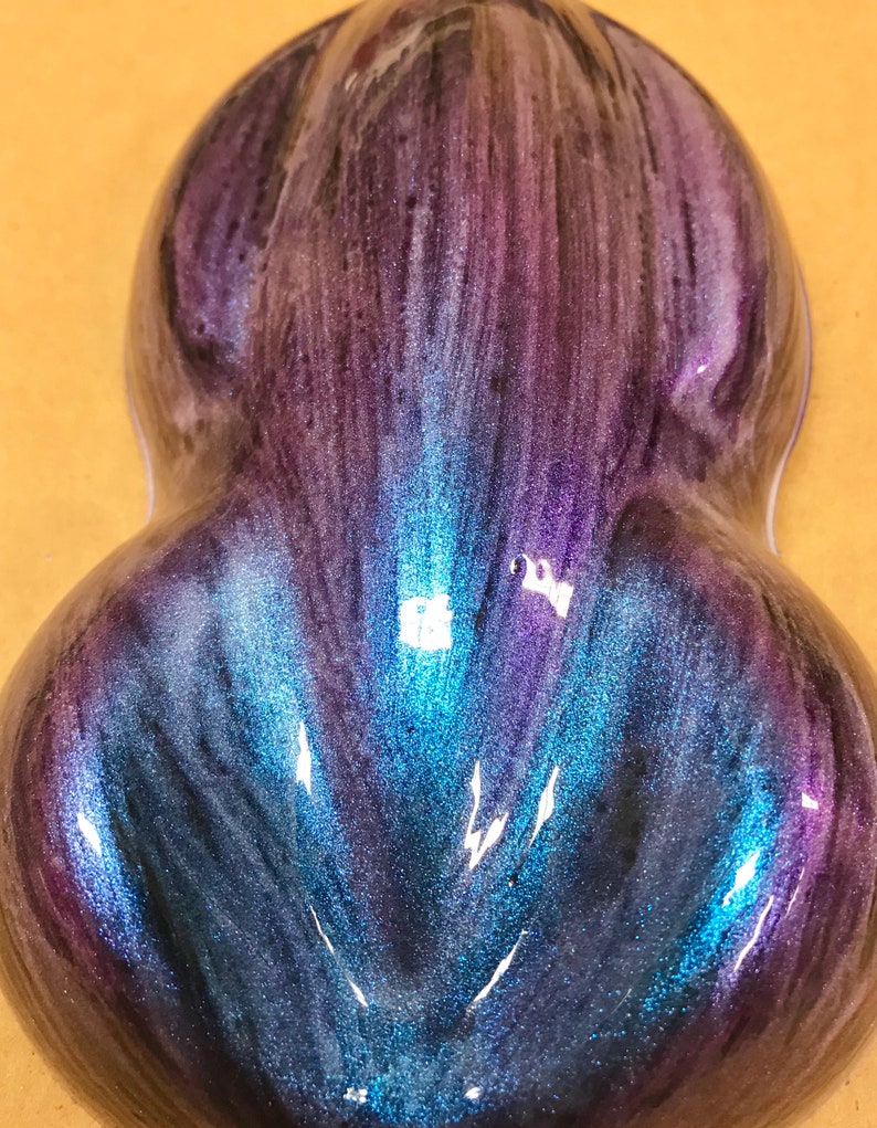 May include: A teardrop-shaped object with a shimmering, iridescent finish. The colours shift from deep purple to vibrant blue and teal, creating a glossy effect. The surface reflects light, highlighting the texture. The object is approximately 10 cm in length.