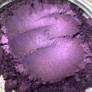 May include: Close-up of a container filled with a shimmering, deep purple powder. The powder has a metallic sheen and appears to have been swirled, creating textured lines across the surface. The color shifts in the light, revealing lighter shades of purple.