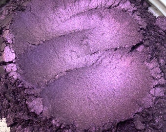 May include: Close-up of a container filled with a shimmering, deep purple powder. The powder has a metallic sheen and appears to have been swirled, creating textured lines across the surface. The color shifts in the light, revealing lighter shades of purple.