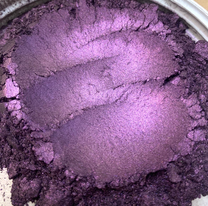 May include: Close-up of a container filled with a shimmering, deep purple powder. The powder has a metallic sheen and appears to have been swirled, creating textured lines across the surface. The color shifts in the light, revealing lighter shades of purple.