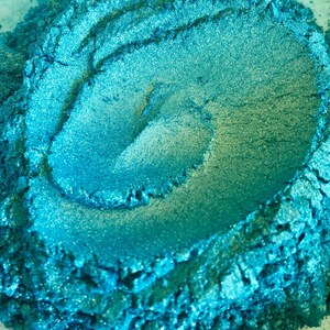 May include: Close-up of a pile of vibrant, shimmering teal powder. The powder has a textured, granular appearance with areas of lighter and darker shades of teal, creating depth and visual interest. The product appears to be a cosmetic pigment or craft material.
