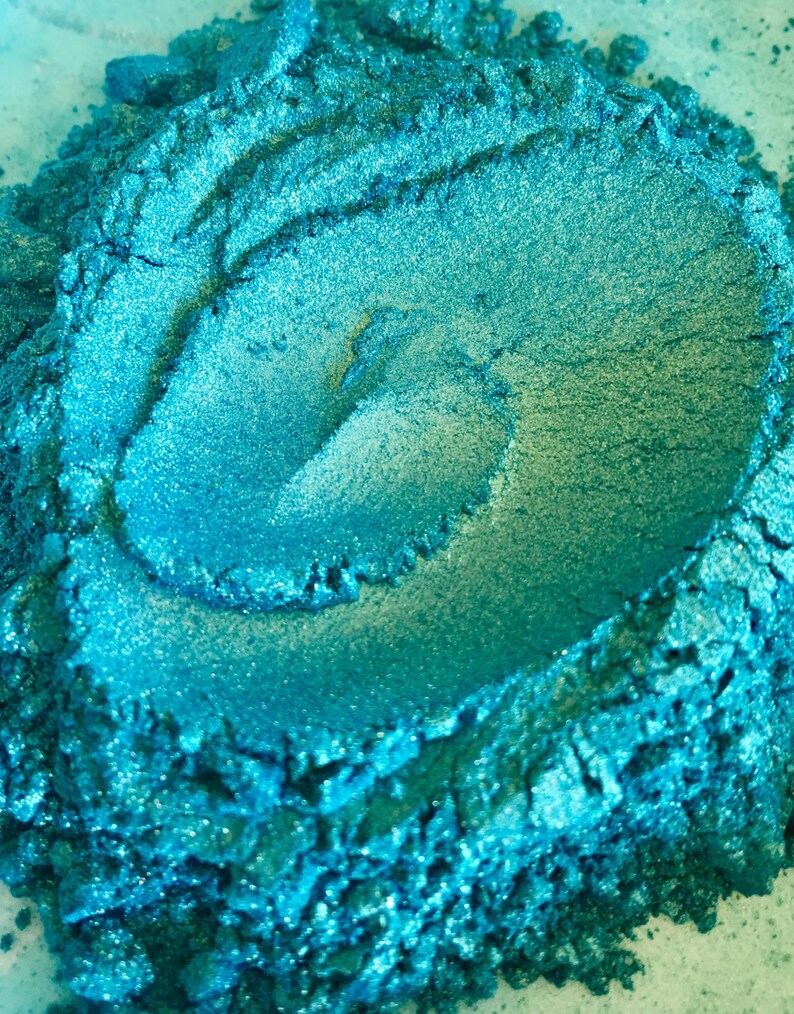May include: Close-up of a pile of vibrant, shimmering teal powder. The powder has a textured, granular appearance with areas of lighter and darker shades of teal, creating depth and visual interest. The product appears to be a cosmetic pigment or craft material.