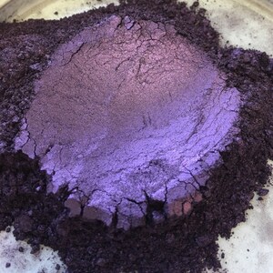 May include: Close-up of a pile of purple and dark purple powder. The powder has a metallic sheen and appears to be finely milled. The texture is slightly cracked, with variations in color and tone. The powder is likely a pigment or cosmetic product.