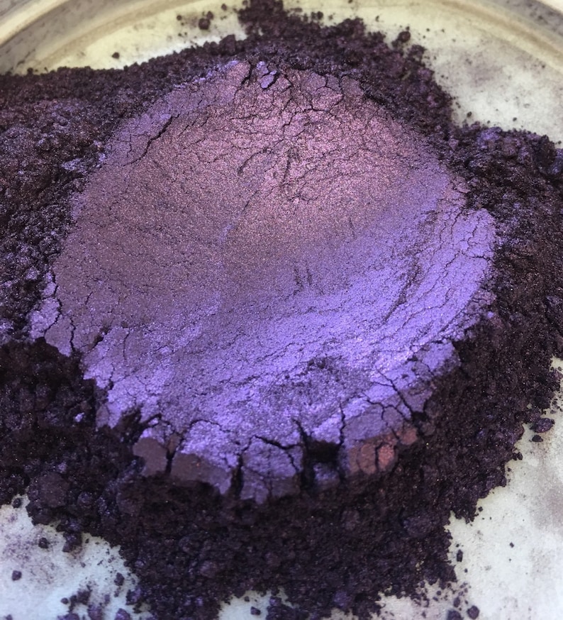 May include: Close-up of a pile of purple and dark purple powder. The powder has a metallic sheen and appears to be finely milled. The texture is slightly cracked, with variations in color and tone. The powder is likely a pigment or cosmetic product.