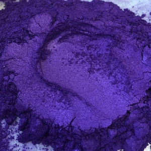 Puede incluir: Primer plano de un mont&oacute;n de polvo de pigmento morado intenso. El polvo tiene un brillo met&aacute;lico, con variaciones de tono y textura. La imagen destaca la naturaleza fina y granular del producto, lo que sugiere su uso en artes y manualidades o cosm&eacute;ticos.