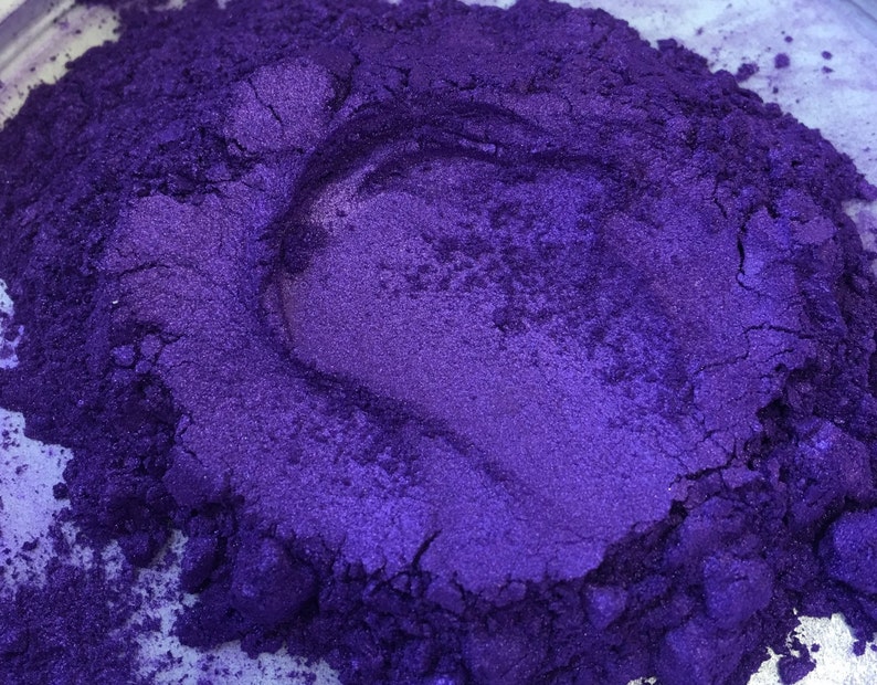 Puede incluir: Primer plano de un mont&oacute;n de polvo de pigmento morado intenso. El polvo tiene un brillo met&aacute;lico, con variaciones de tono y textura. La imagen destaca la naturaleza fina y granular del producto, lo que sugiere su uso en artes y manualidades o cosm&eacute;ticos.