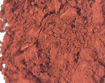 May include: Close-up of a pile of fine, reddish-brown powder. The powder has a textured, granular appearance with varying shades of red and brown. The background is white, providing contrast to the powder.