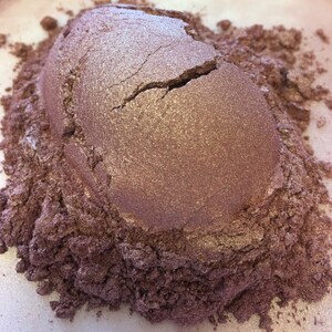 May include: A close-up of a pile of shimmering, metallic-looking powder in shades of mauve and bronze. The powder has a cracked, textured surface, with lighter highlights catching the light. The surrounding powder is a darker, granular texture.