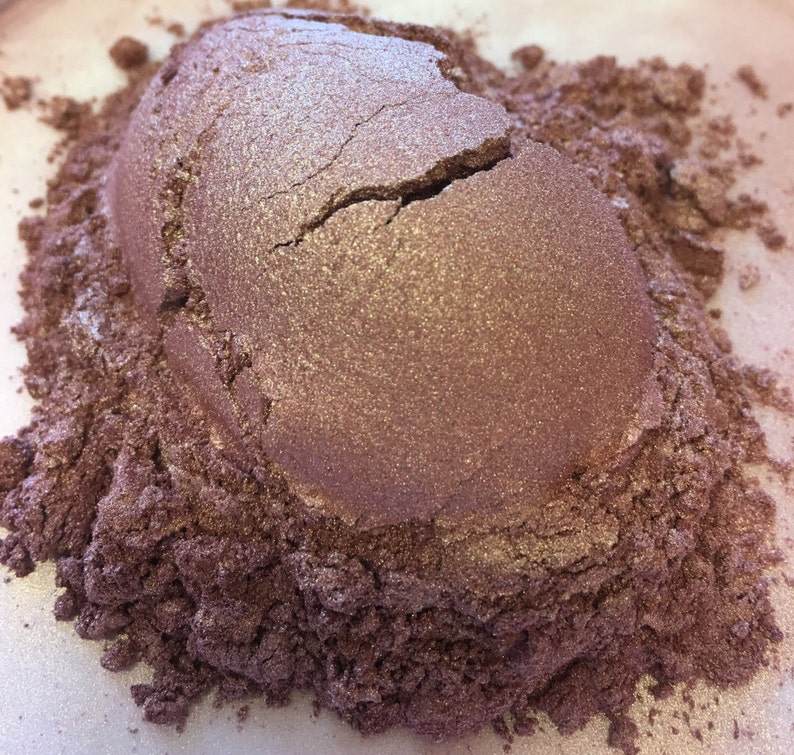 May include: A close-up of a pile of shimmering, metallic-looking powder in shades of mauve and bronze. The powder has a cracked, textured surface, with lighter highlights catching the light. The surrounding powder is a darker, granular texture.