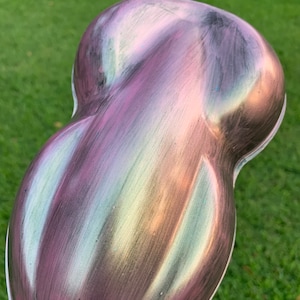 May include: A pearlescent, iridescent object with a smooth, curved shape. The surface transitions through shades of purple, green, and gold, creating a shimmering effect. The object is set against a backdrop of green grass.