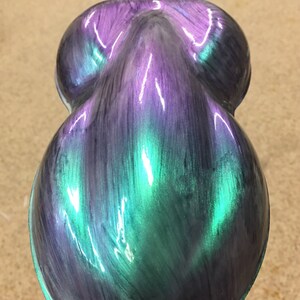 Puede incluir: Un objeto nacarado en forma de l&aacute;grima con un acabado que cambia de color. La superficie transita del morado al verde azulado y al verde, con un brillo brillante. La forma del objeto es suave y redondeada, lo que sugiere una pieza decorativa o art&iacute;stica.