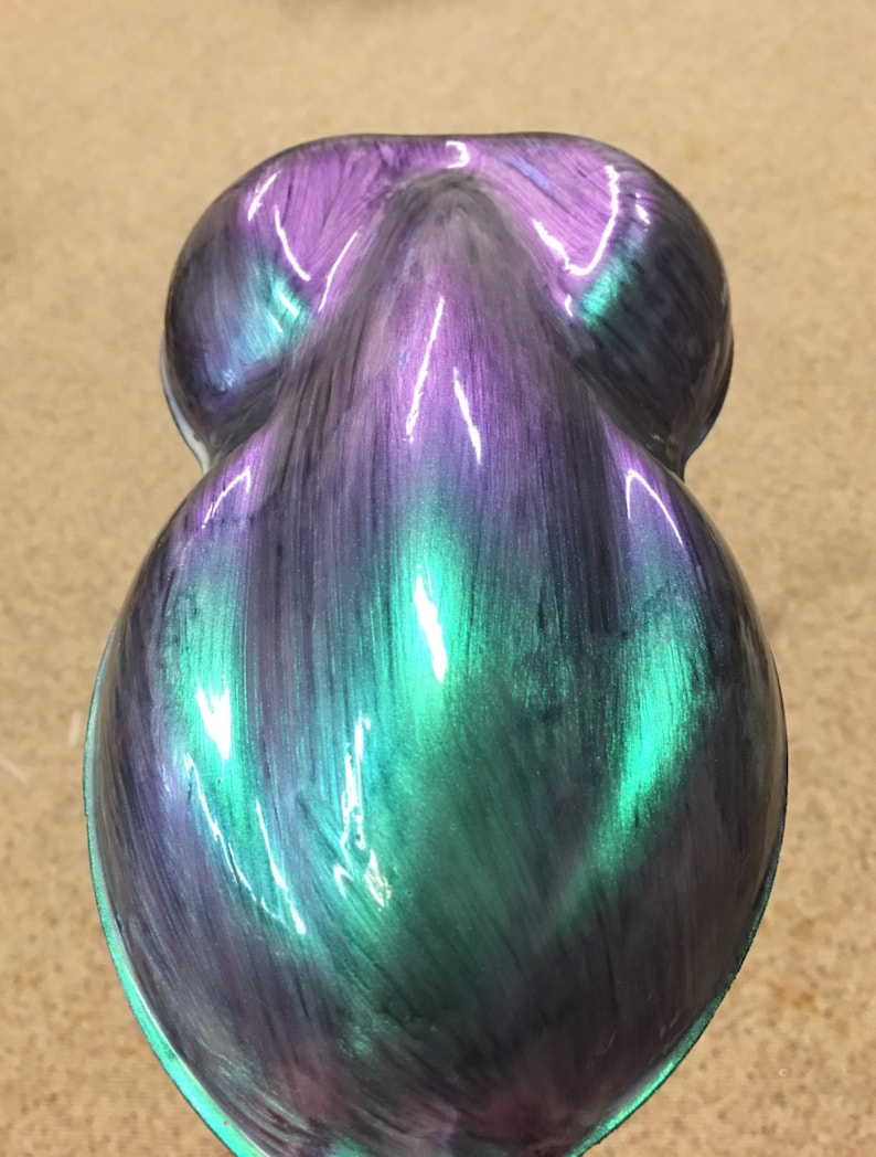 Puede incluir: Un objeto nacarado en forma de l&aacute;grima con un acabado que cambia de color. La superficie transita del morado al verde azulado y al verde, con un brillo brillante. La forma del objeto es suave y redondeada, lo que sugiere una pieza decorativa o art&iacute;stica.