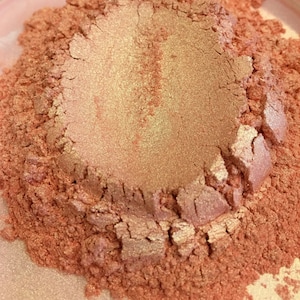 May include: Close-up of a pile of shimmering, finely milled, rose-gold colored powder. The powder has a textured, granular appearance with areas of light reflection, suggesting a cosmetic product like eyeshadow or highlighter.