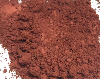 May include: Close-up of a pile of fine, reddish-brown powder. The powder has a textured, granular appearance, with varying shades of brown. The image is well-lit, highlighting the details of the powder's texture. The powder appears to be a pigment or dye.