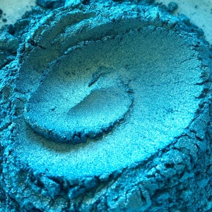 May include: Close-up of a pile of shimmering, teal-colored powder. The powder has a metallic sheen and a textured surface, with visible ridges and a swirling pattern. The color shifts slightly with the light, creating highlights and shadows.