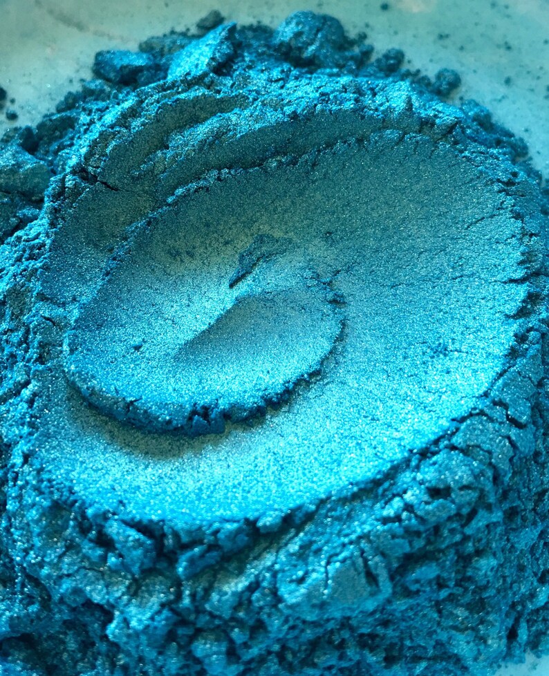 May include: Close-up of a pile of shimmering, teal-colored powder. The powder has a metallic sheen and a textured surface, with visible ridges and a swirling pattern. The color shifts slightly with the light, creating highlights and shadows.