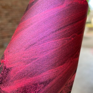 May include: Close-up of a rolled sheet of material with a vibrant, swirling pattern. The colors transition from deep burgundy to bright magenta, with hints of black. The texture appears slightly metallic.
