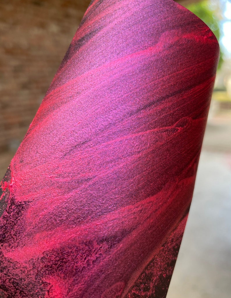 May include: Close-up of a rolled sheet of material with a vibrant, swirling pattern. The colors transition from deep burgundy to bright magenta, with hints of black. The texture appears slightly metallic.