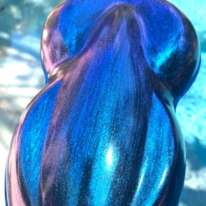 May include: A pearlescent, teardrop-shaped object with a glossy finish. The surface transitions from deep blue to purple, with streaks of teal and iridescent shimmer. The object's form is smooth and rounded, reflecting light.