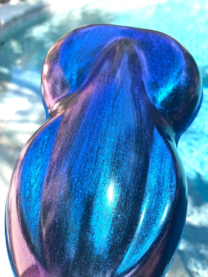 May include: A pearlescent, teardrop-shaped object with a glossy finish. The surface transitions from deep blue to purple, with streaks of teal and iridescent shimmer. The object's form is smooth and rounded, reflecting light.