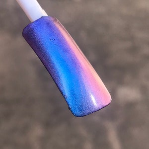 May include: A nail swatch displays a gradient of iridescent colors. The colors transition from a deep blue at the base to a soft pink at the tip, with a vibrant blue in the middle. The nail polish has a shimmering, metallic finish.