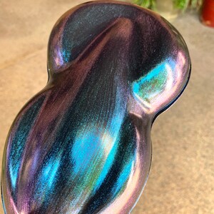 May include: A pearlescent, iridescent paint sample with a unique, organic shape. The color shifts from deep purple to teal and blue, with hints of pink and gold. The surface has a glossy finish, reflecting light and creating a shimmering effect.