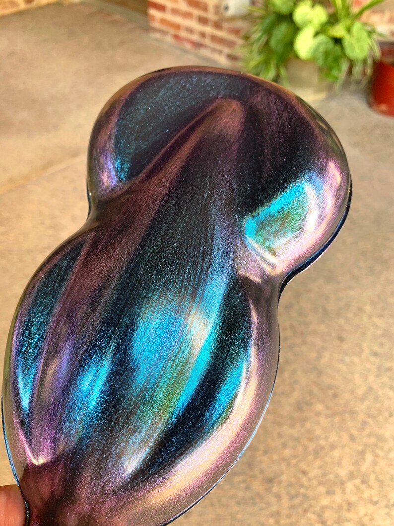 May include: A pearlescent, iridescent paint sample with a unique, organic shape. The color shifts from deep purple to teal and blue, with hints of pink and gold. The surface has a glossy finish, reflecting light and creating a shimmering effect.
