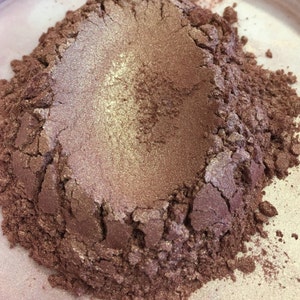 May include: Close-up of a pile of shimmering, metallic-looking powder. The powder has a gradient of colors, with a light gold center and a darker, reddish-brown hue around the edges. The texture appears granular and slightly uneven.