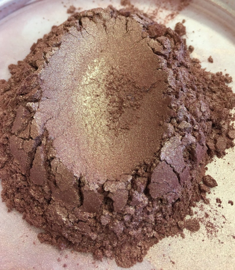 May include: Close-up of a pile of shimmering, metallic-looking powder. The powder has a gradient of colors, with a light gold center and a darker, reddish-brown hue around the edges. The texture appears granular and slightly uneven.