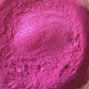 May include: Close-up of a vibrant, textured, magenta-colored powder. The powder has a shimmering, metallic sheen, suggesting it may be a cosmetic pigment or craft material. The surface is uneven, with subtle variations in color and texture.