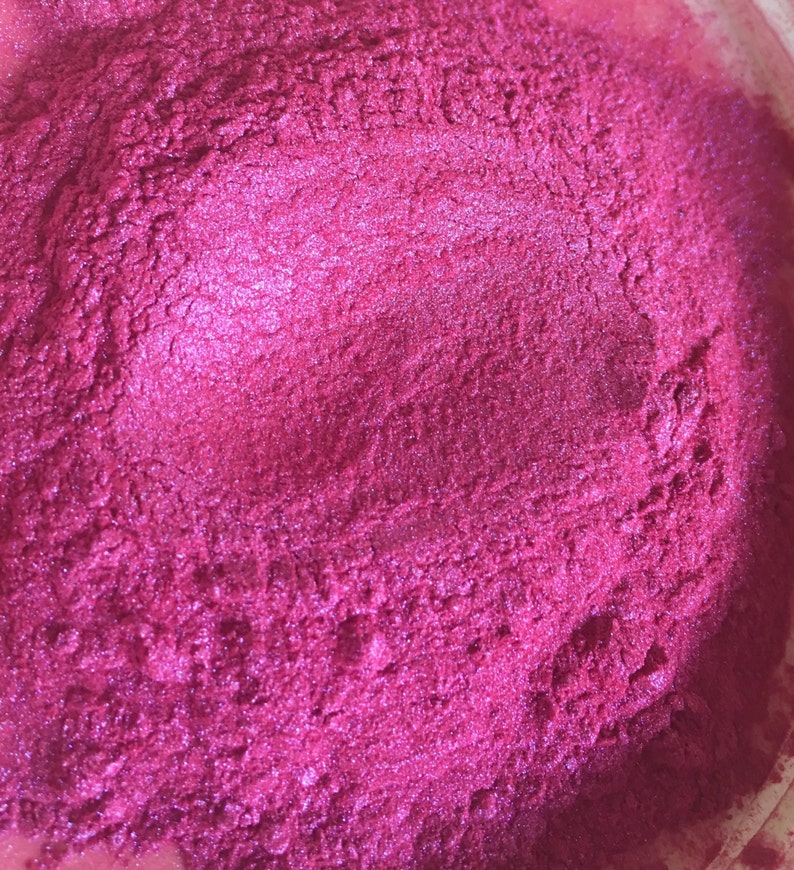 May include: Close-up of a vibrant, textured, magenta-colored powder. The powder has a shimmering, metallic sheen, suggesting it may be a cosmetic pigment or craft material. The surface is uneven, with subtle variations in color and texture.