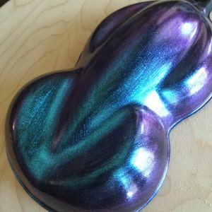 May include: A pearlescent, iridescent paint sample in a teardrop shape. The color shifts between shades of purple, teal, and blue, with a glossy finish. The sample is displayed on a light-colored wooden surface.