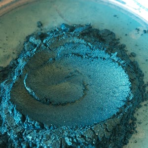 May include: Close-up of a container filled with teal-colored pigment powder. The powder has a shimmering, metallic finish and a textured appearance. The container is white with a teal interior.